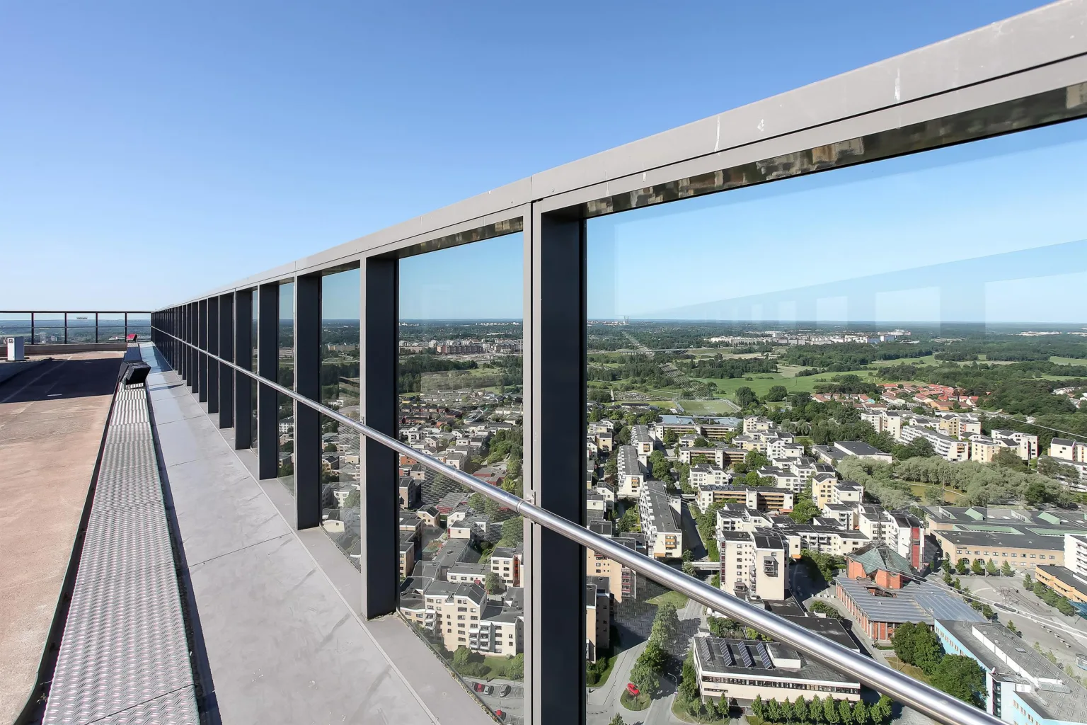 En hög balkong med glasbarriärer som ger en panoramautsikt över en stad med grönområden och byggnader.