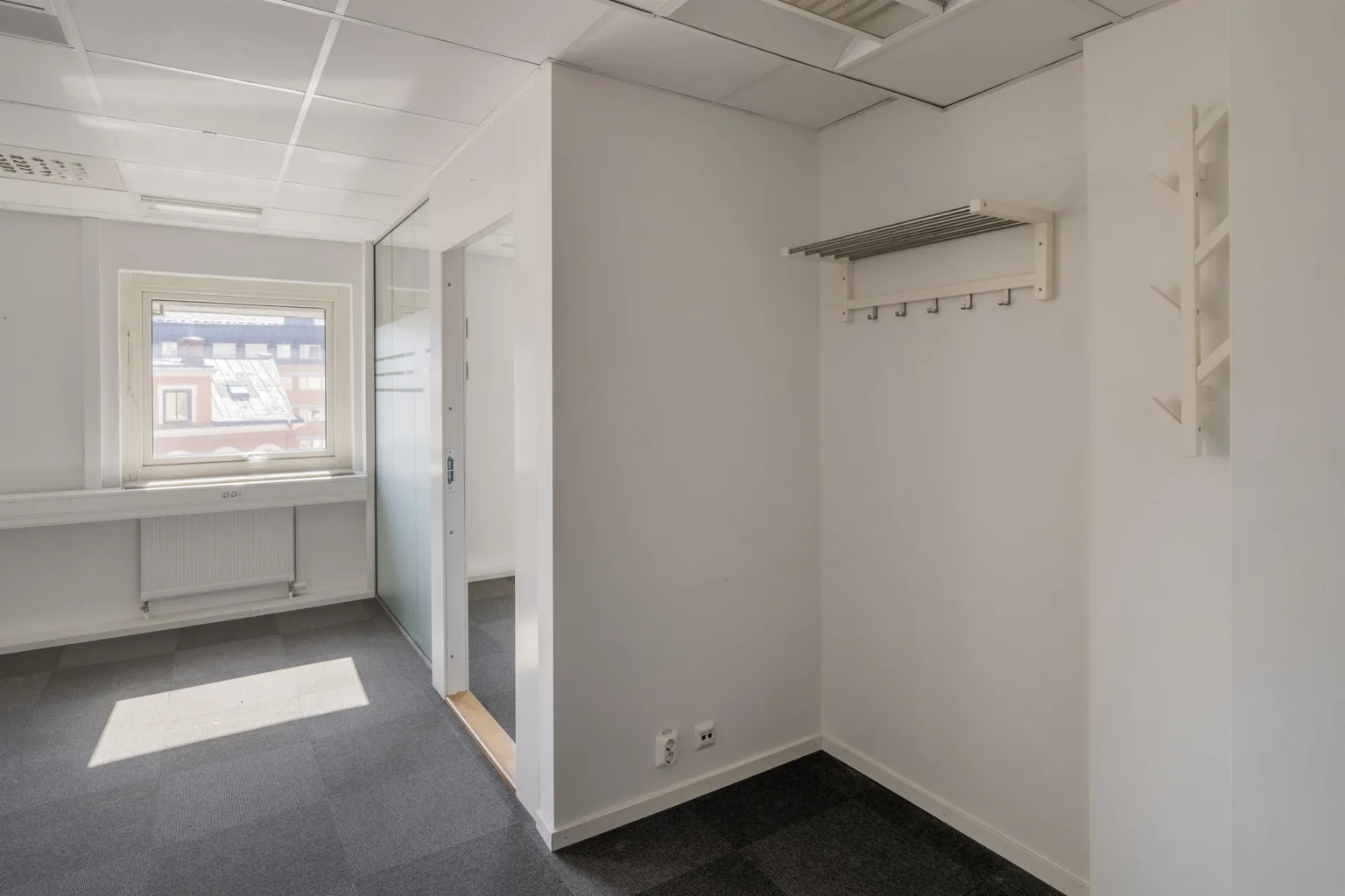 En vit hall med klädhängare på väggen och fönster i bakgrunden.