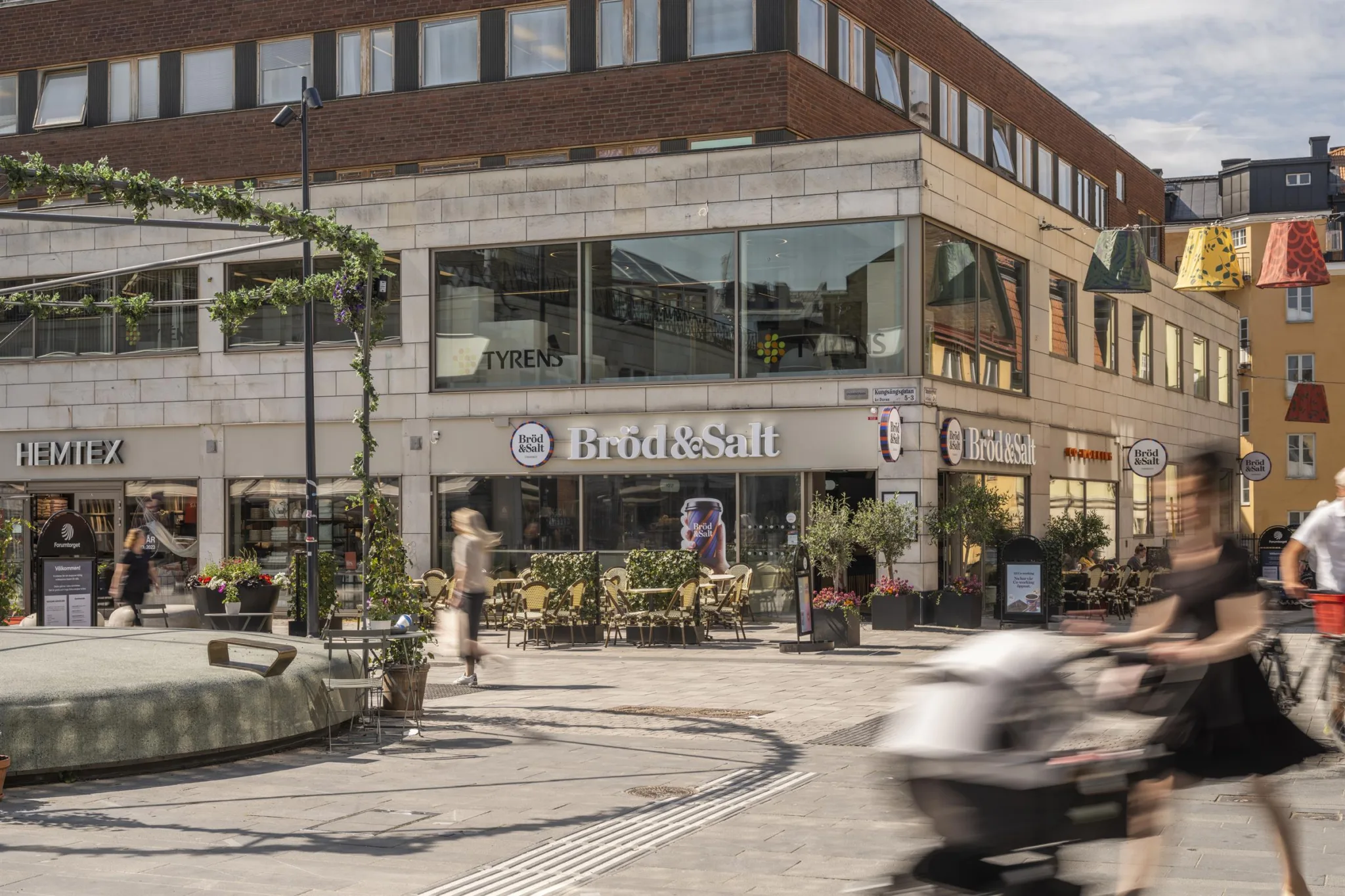 En byggnad med en butiksskylt där det står "Friskis," omgiven av uteservering och människor.