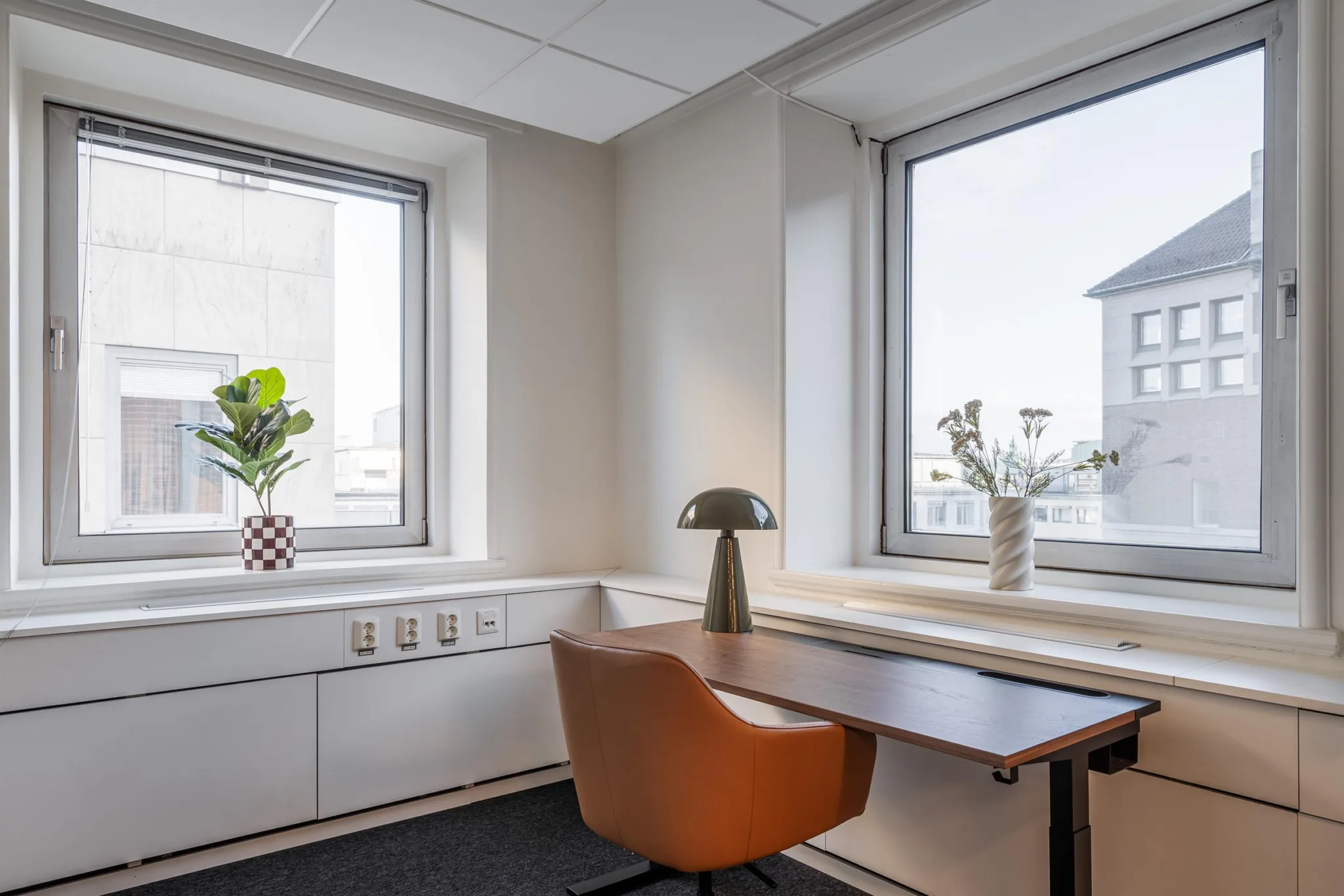 2. Ett skrivbord med en orange stol, en svart bordslampa och växter placerade vid två stora fönster i ett ljust kontorsutrymme.