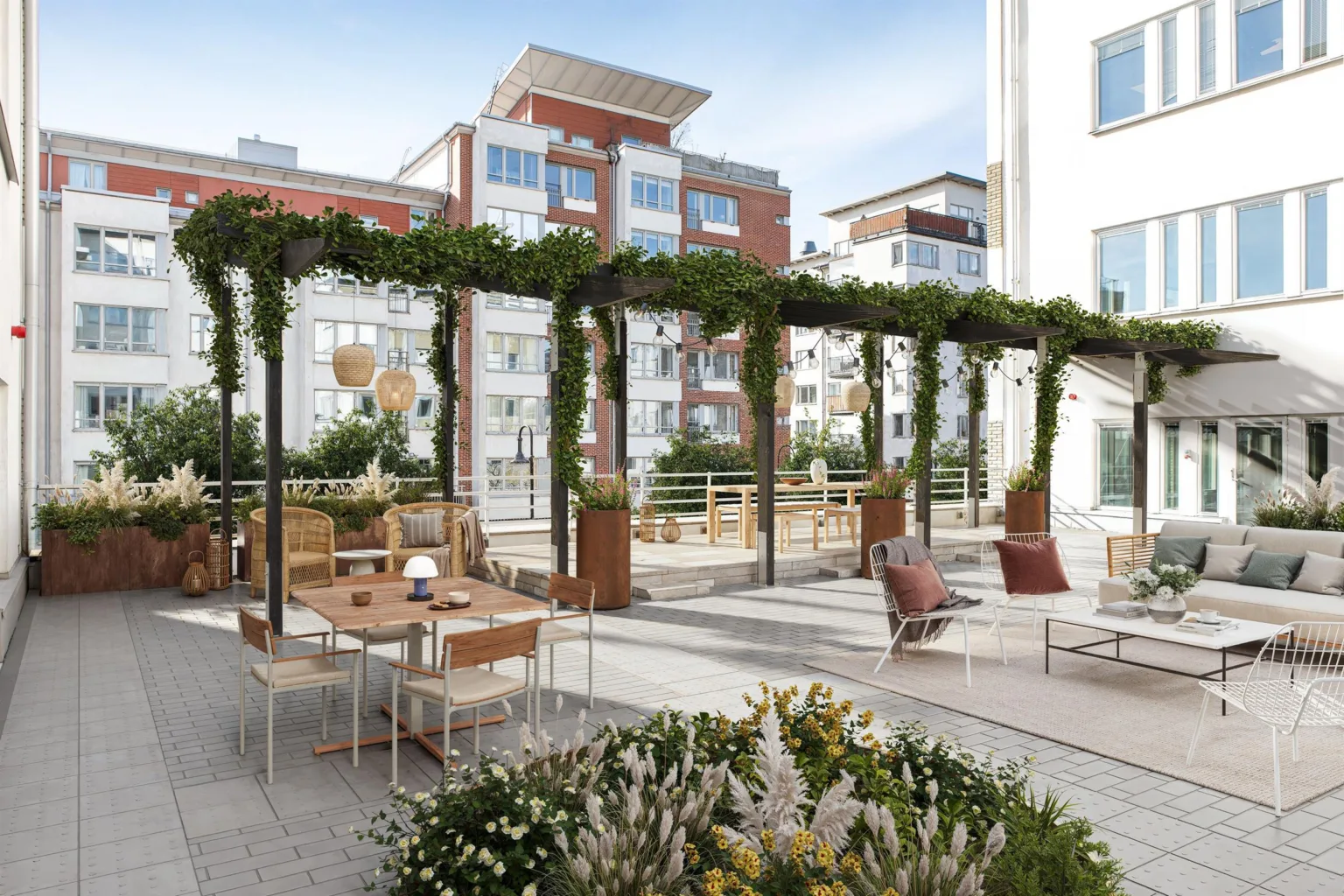 En takterrass med möbler, växter och pergola täckt med grönska, med utsikt över omgivande byggnader.