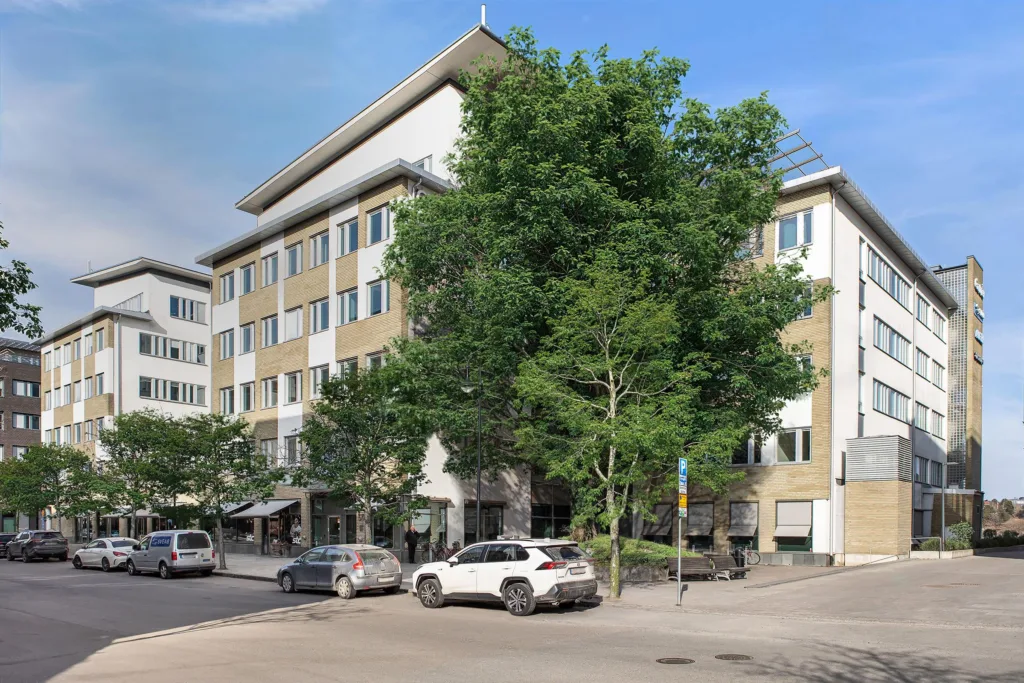 En flerbostadsbyggnad med vita och gula fasader, träd framför och bilar parkerade vid trottoaren.
