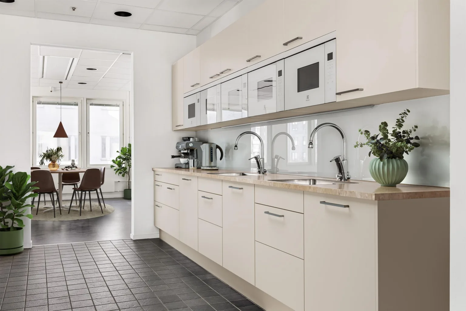 5. Ett modernt kök med ljusa skåp, flera kranar, och en kaffemaskin, med ett matbord synligt i bakgrunden.