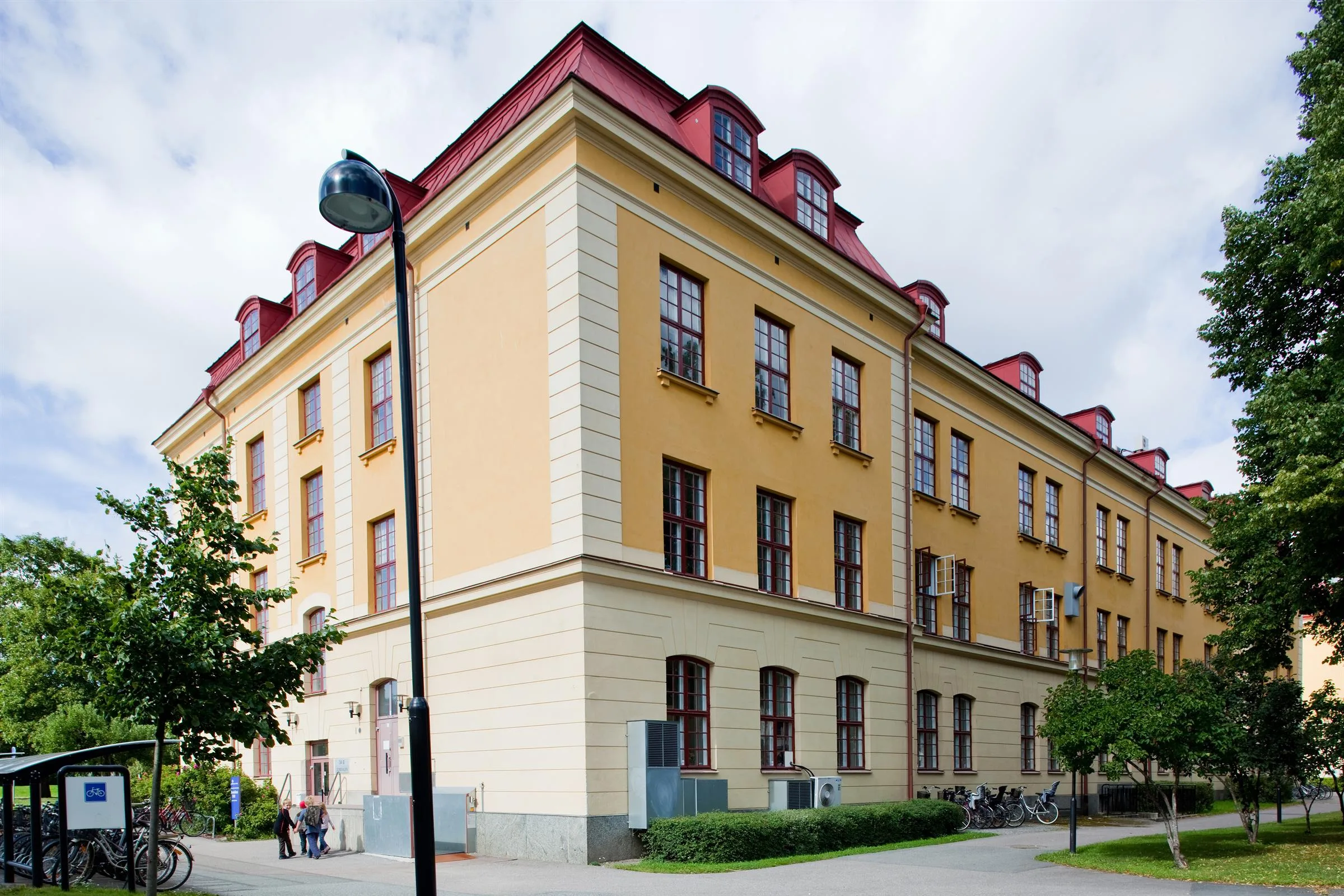 1. En gul byggnad med röda takfönster och flera stora fönster, omgiven av grönska.