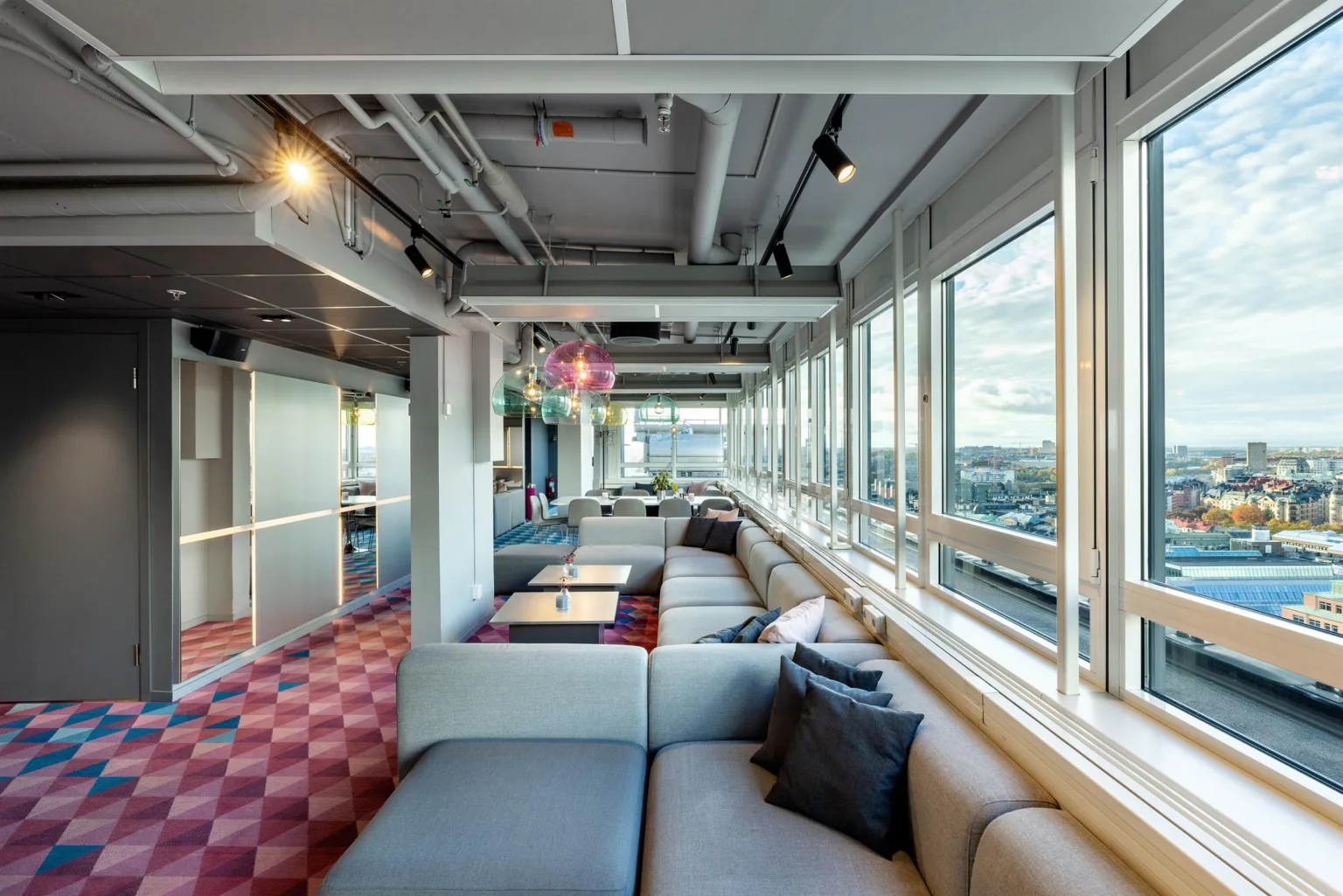 1. En lounge med grå soffor och kuddar i ett rum med stora fönster som ger utsikt över staden.