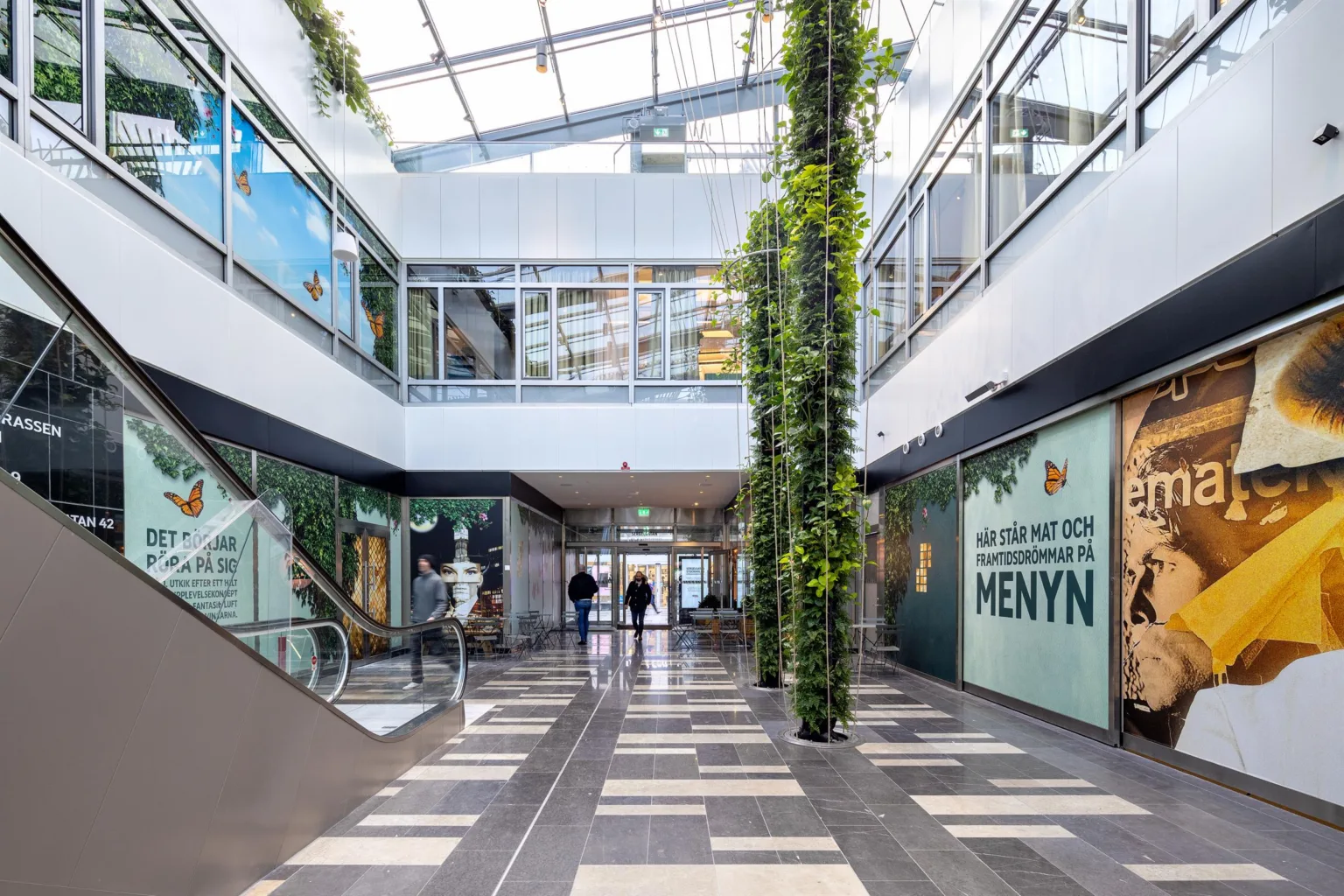 En ljus inomhusgalleria med växter, glasväggar och skylten "MENYN".
