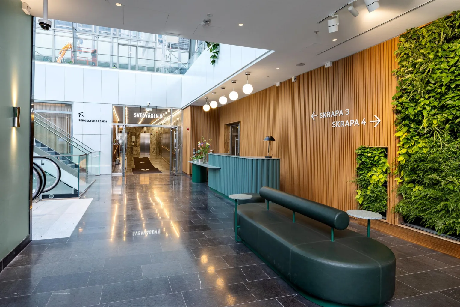 En reception med träpaneler, gröna växter och en grön sittmöbel i en ljus och modern interiör.