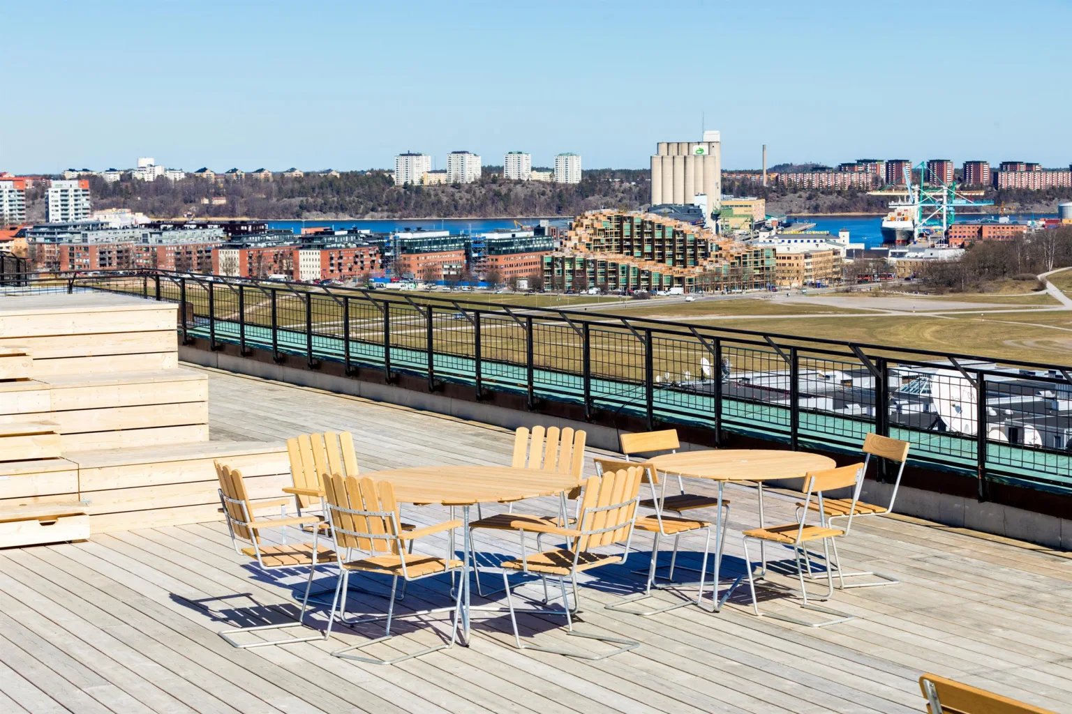 En takterrass med träbord och stolar, samt utsikt över staden och vatten.