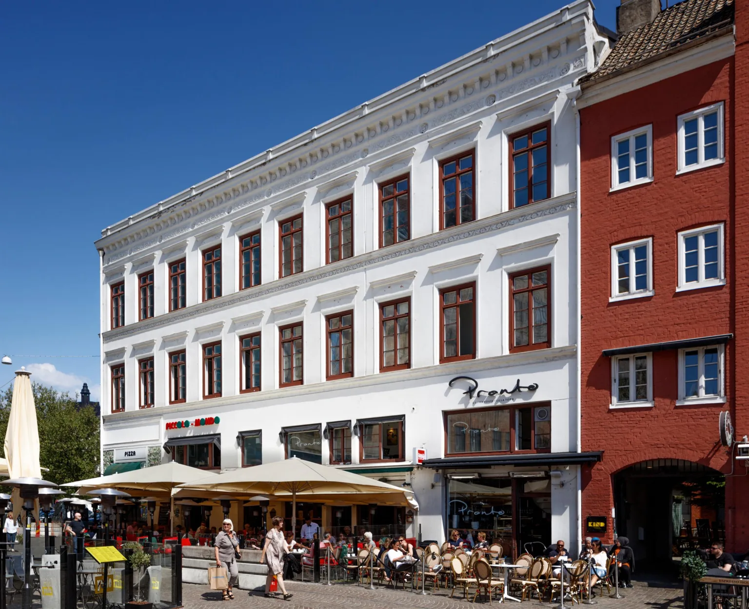 En vit byggnad med stora fönster och uteserveringar framför på en livlig gata.