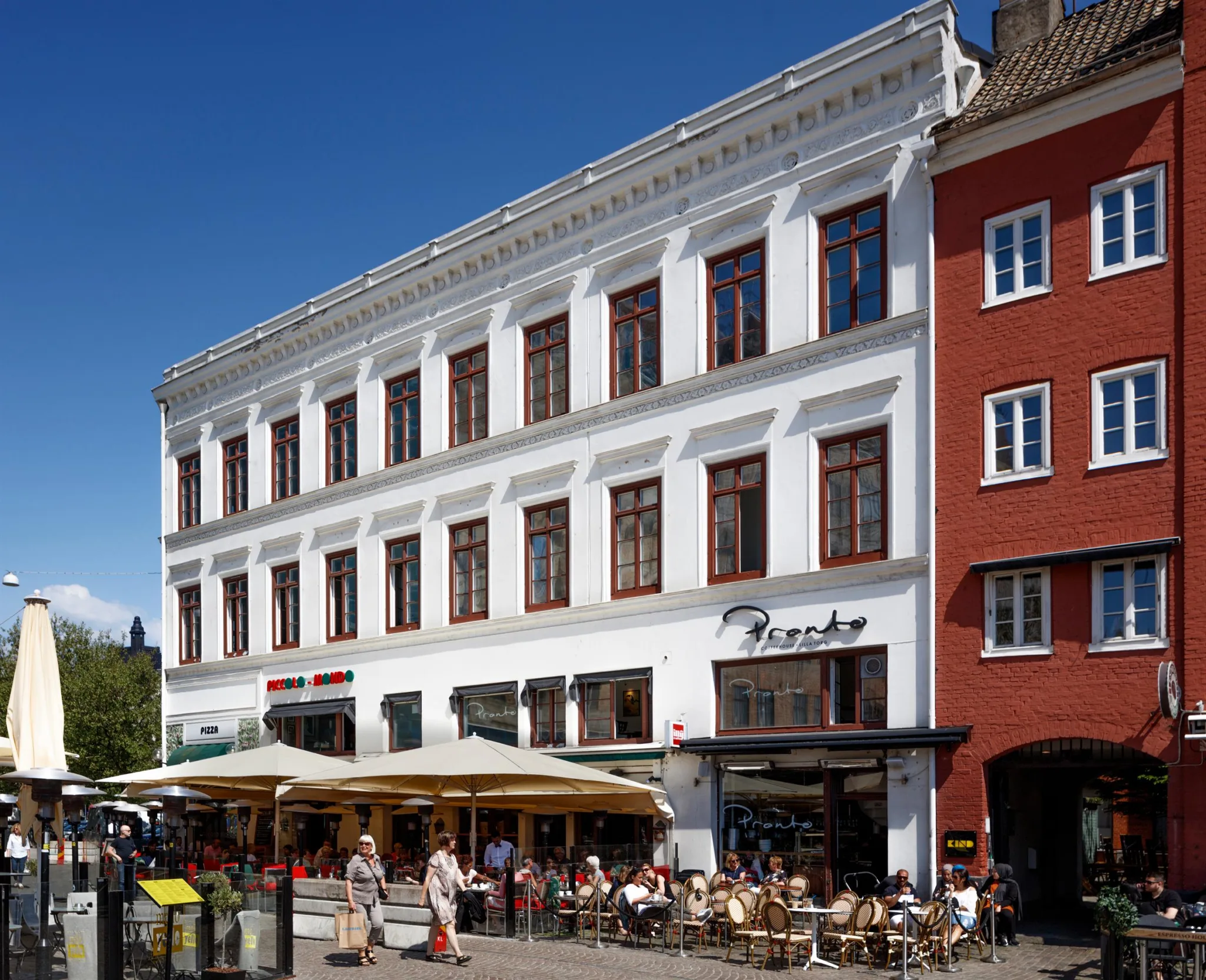 En vit byggnad med stora fönster och uteserveringar framför på en livlig gata.