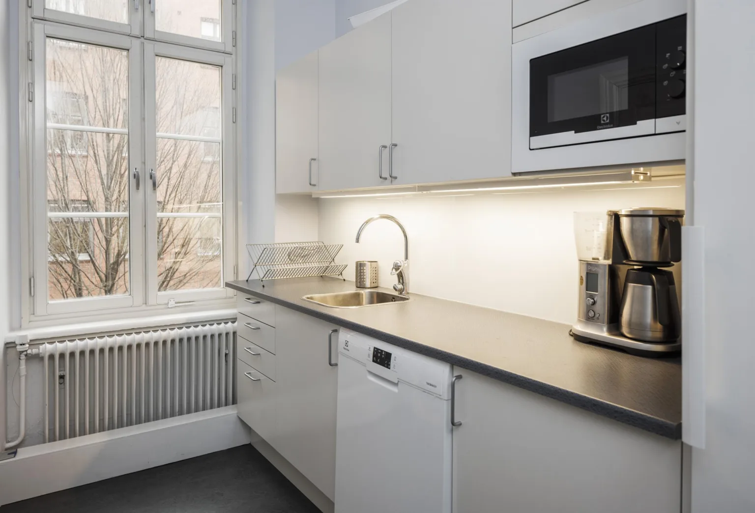 5. Ett kök med vita skåp, bänkskiva, mikrovågsugn, brödrost och ett fönster som släpper in ljus.