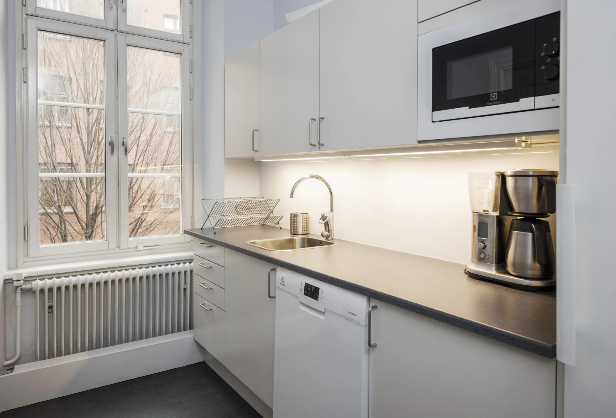 5. Ett kök med vita skåp, bänkskiva, mikrovågsugn, brödrost och ett fönster som släpper in ljus.