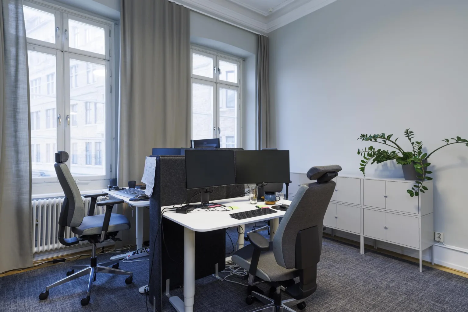 2. Ett kontorsrum med skrivbord, datorskärmar, kontorsstolar och stora fönster med gardiner.