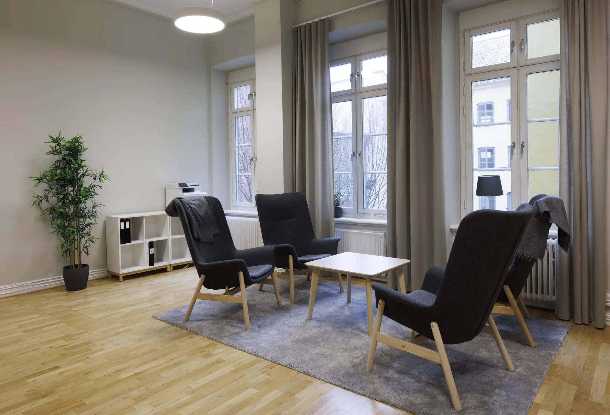 2. Ett möblerat vardagsrum med svarta fåtöljer, ett litet bord på en matta, och stora fönster med gardiner.