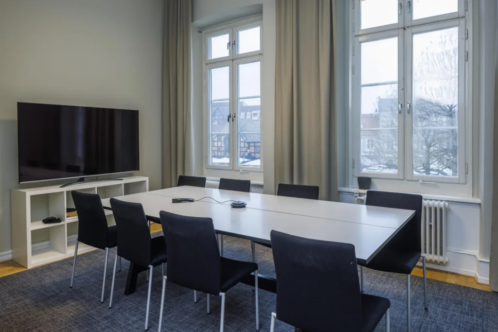 Ett konferensrum med ett långt bord, svarta stolar, stora fönster och en TV.