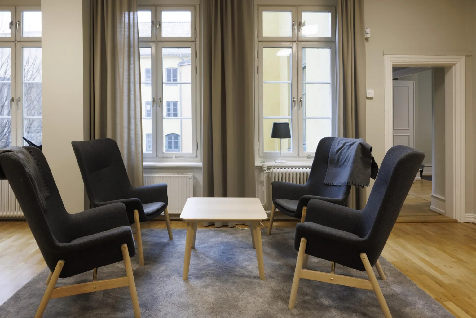 3. Ett mötesrum med fyra fåtöljer i en cirkel runt ett litet bord, omgiven av fönster med gardiner.
