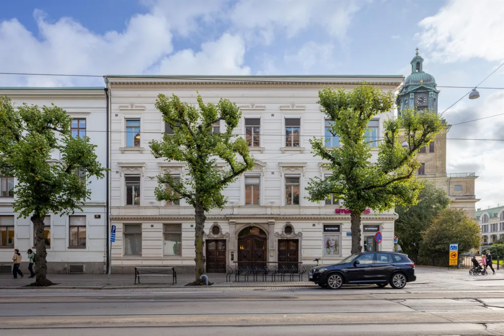 En vit byggnad med tre våningar och valvformad entré, omgiven av träd och en parkerad bil på gatan.