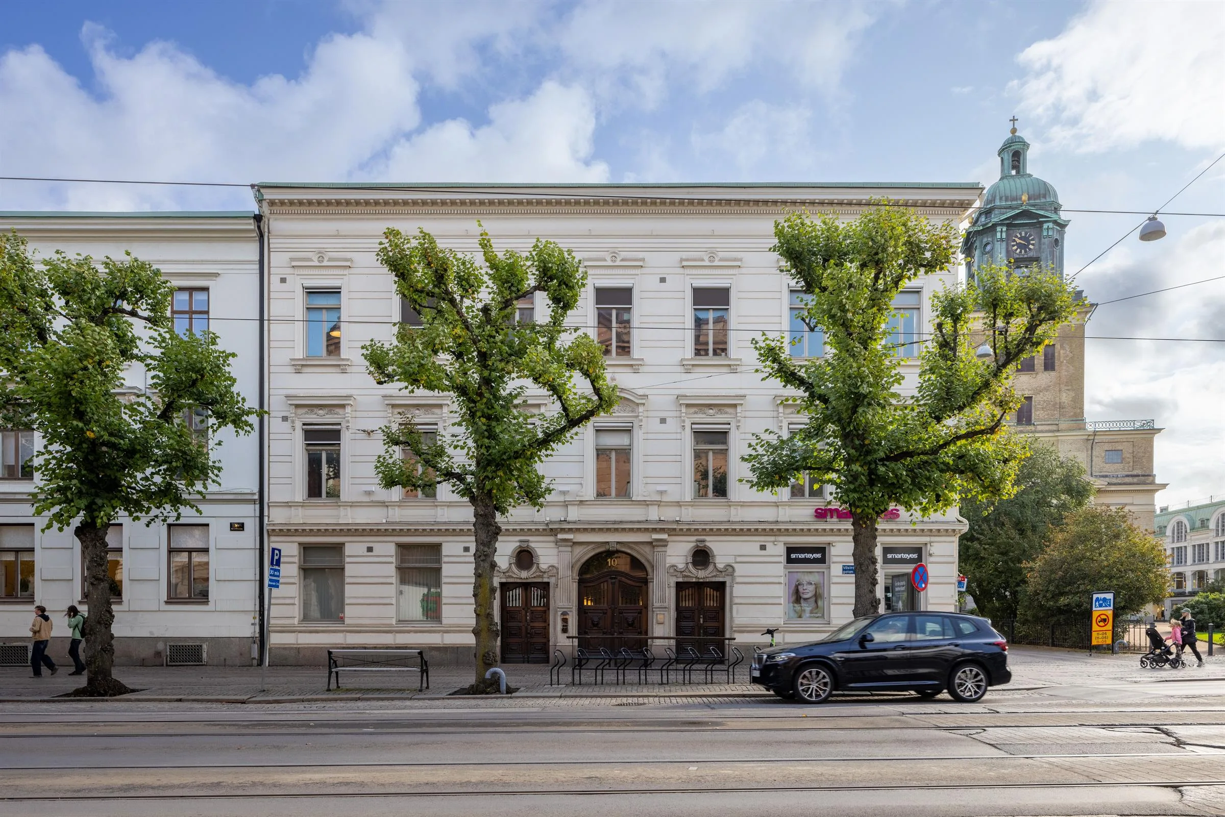 En vit byggnad med tre våningar och valvformad entré, omgiven av träd och en parkerad bil på gatan.