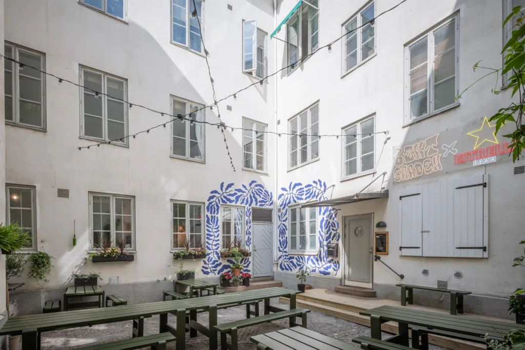 Innergård med vita byggnader, fönster, dekorerad entré med blå mönster och bord med bänkar under ljusslingor.