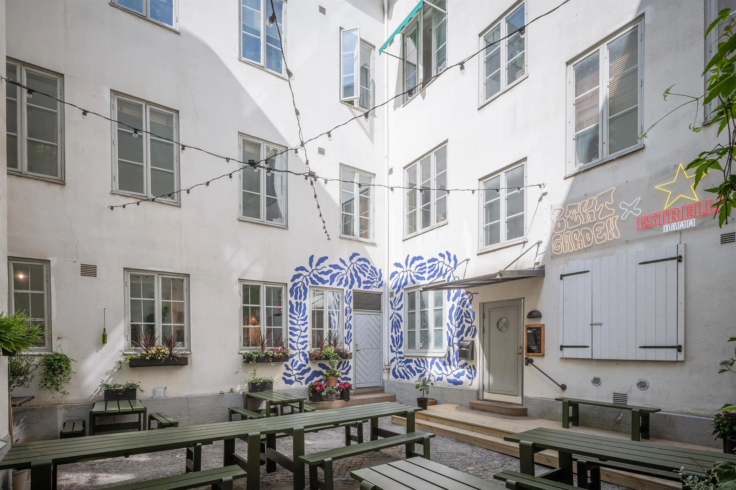 Innergård med vita byggnader, fönster, dekorerad entré med blå mönster och bord med bänkar under ljusslingor.