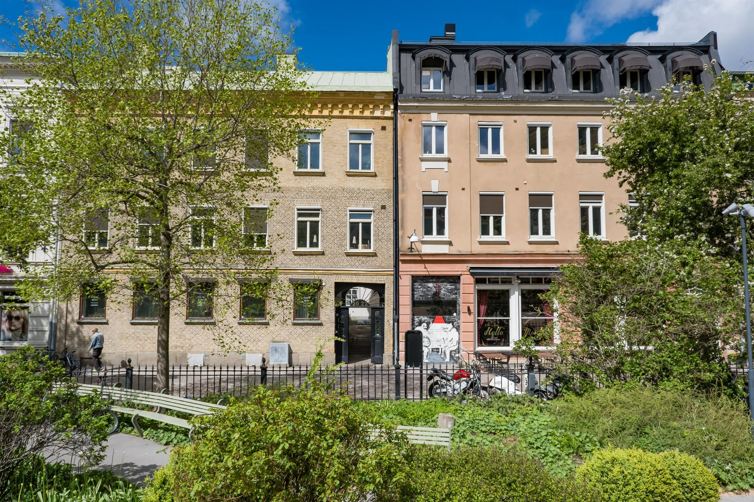 Två byggnader bredvid varandra, en med gul fasad, den andra med beige, omgiven av grönska och cyklar framför entrén.