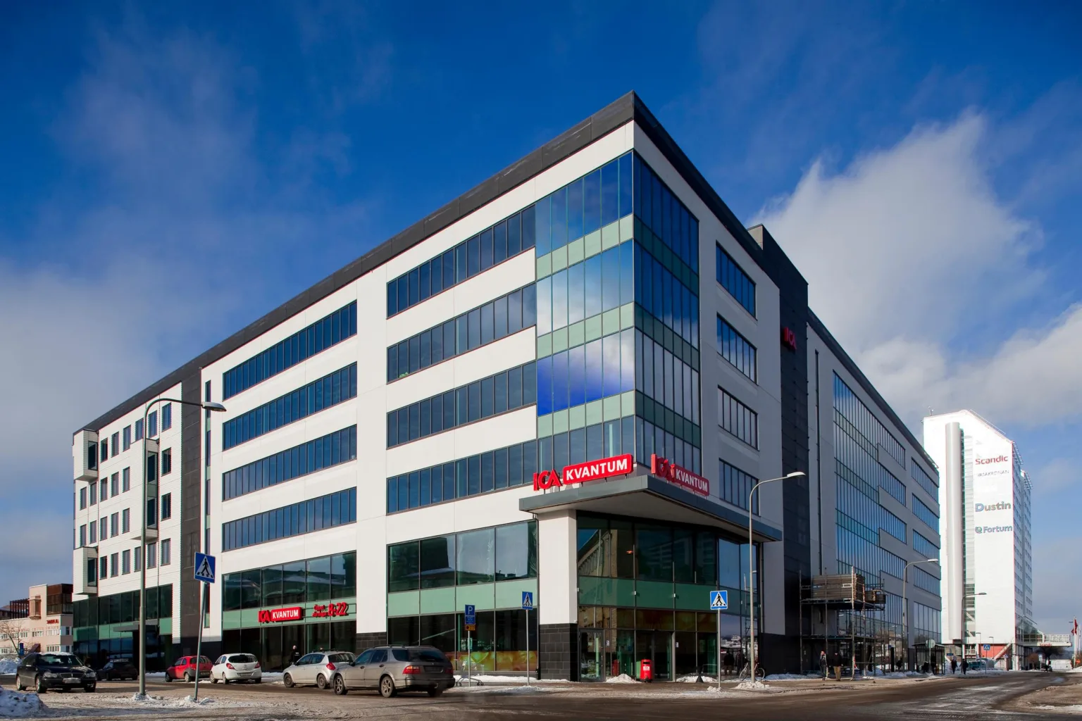En stor modern byggnad med glasfasad, flera våningar och parkerade bilar vid entrén.