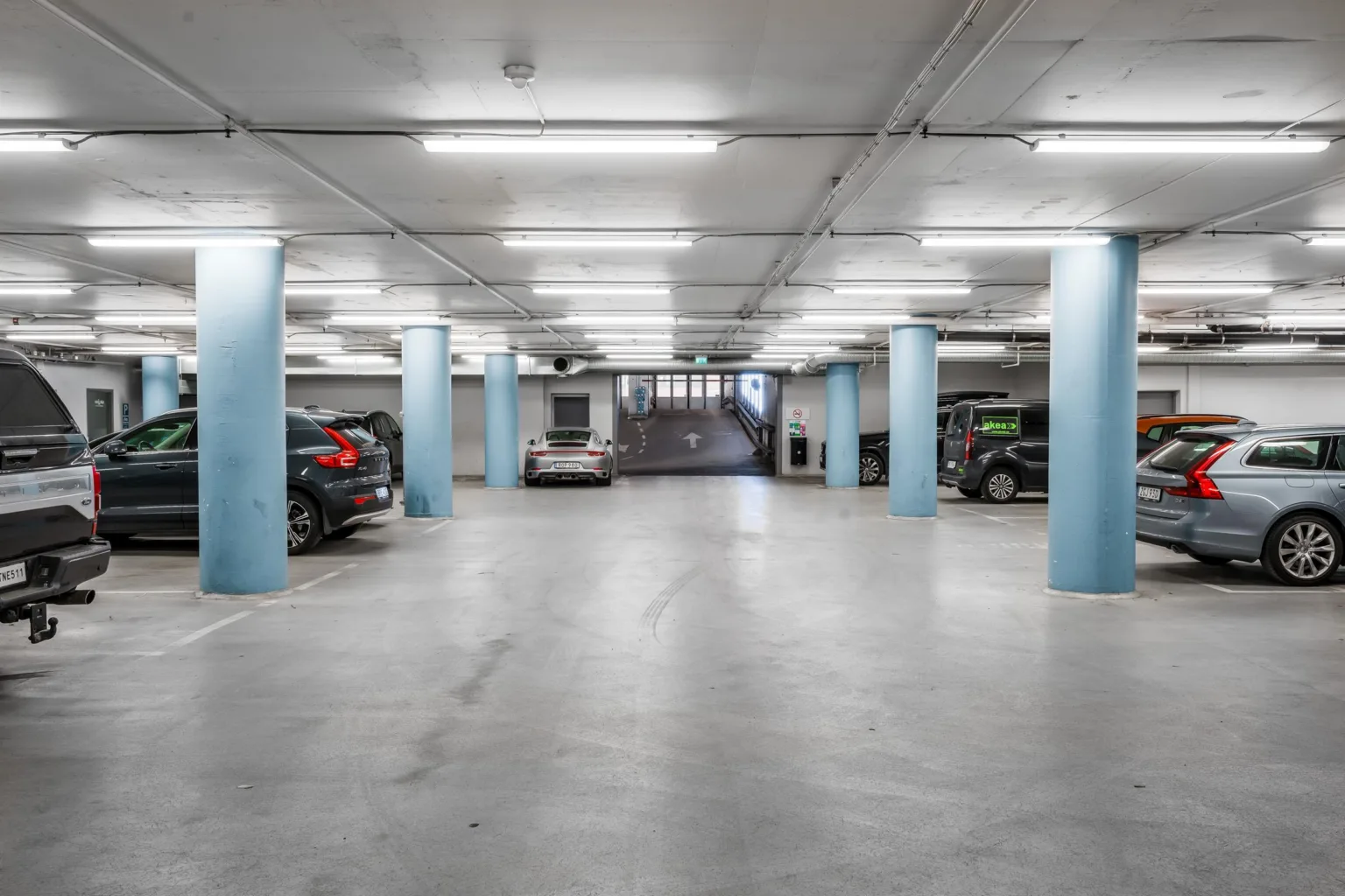 En underjordisk parkeringsplats med blåa pelare, parkerade bilar och stark belysning.
