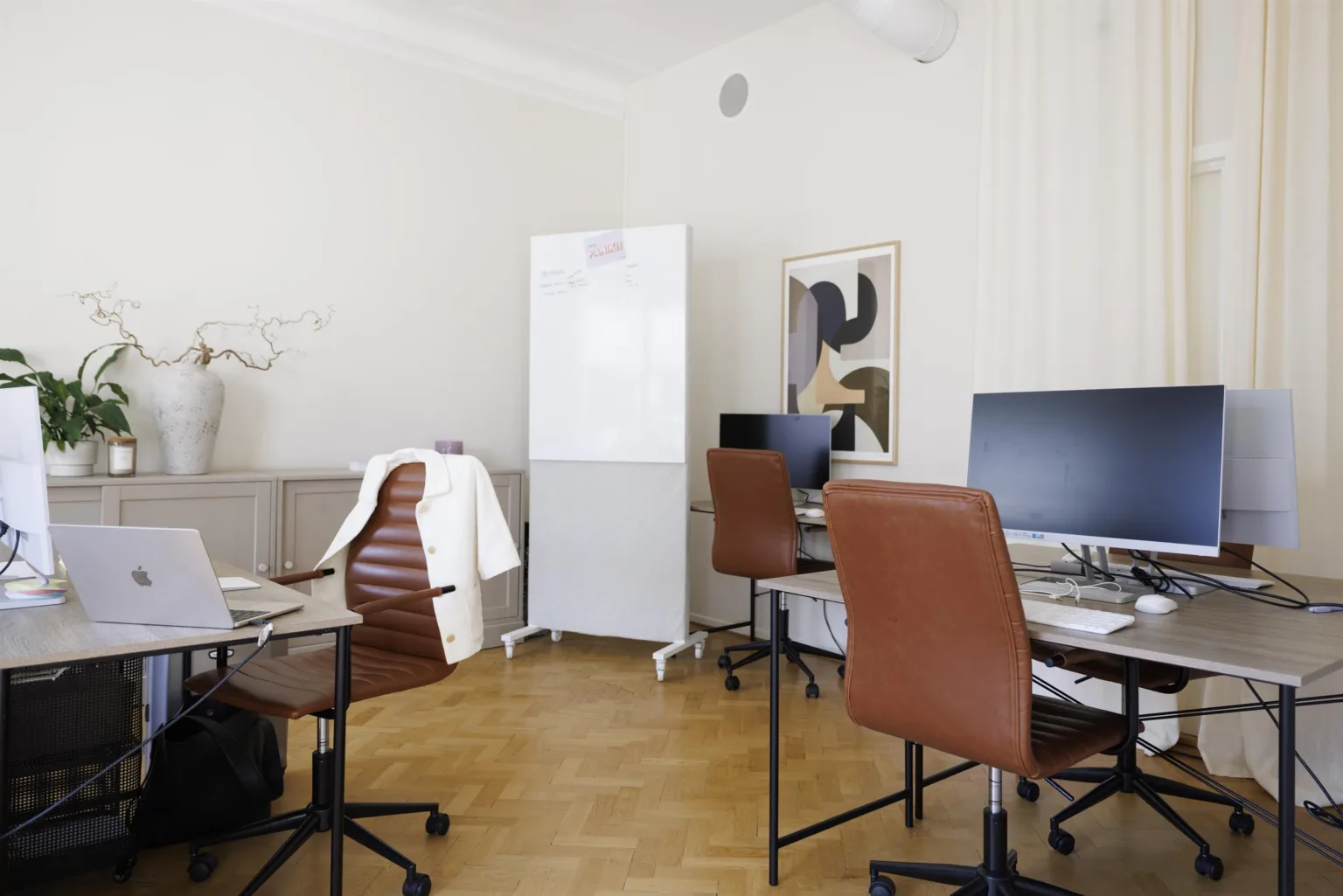 3. Ett kontorsrum med skrivbord, datorer, bruna stolar och en vit förvaringsskåp, dekorerat med en tavla på väggen.