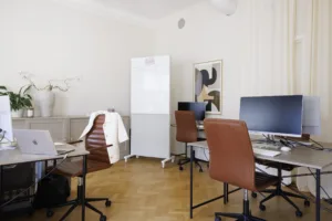 3. Ett kontorsrum med skrivbord, datorer, bruna stolar och en vit förvaringsskåp, dekorerat med en tavla på väggen.