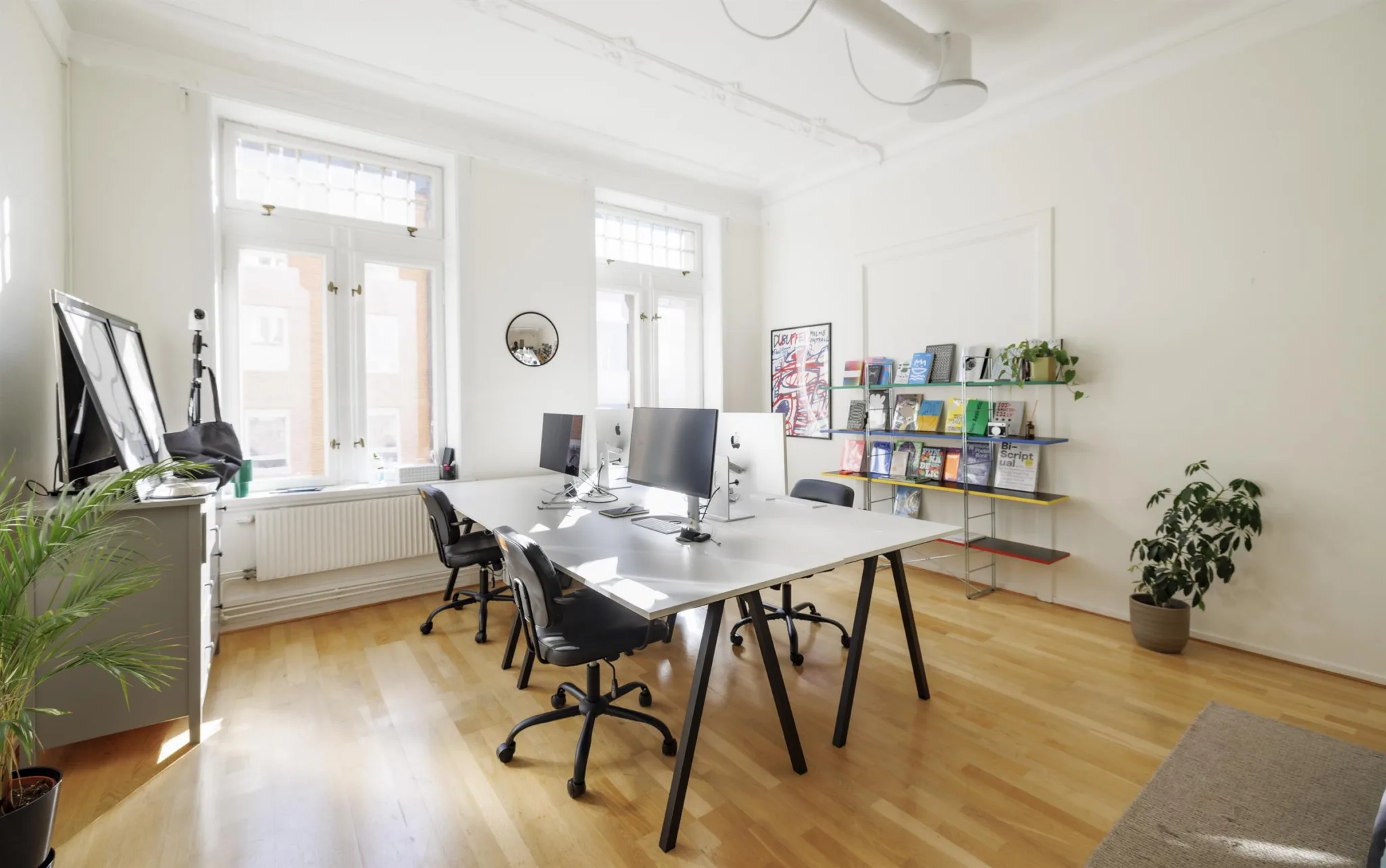 2. Ett ljust kontorsrum med två skrivbord, datorskärmar och hyllor fyllda med böcker.