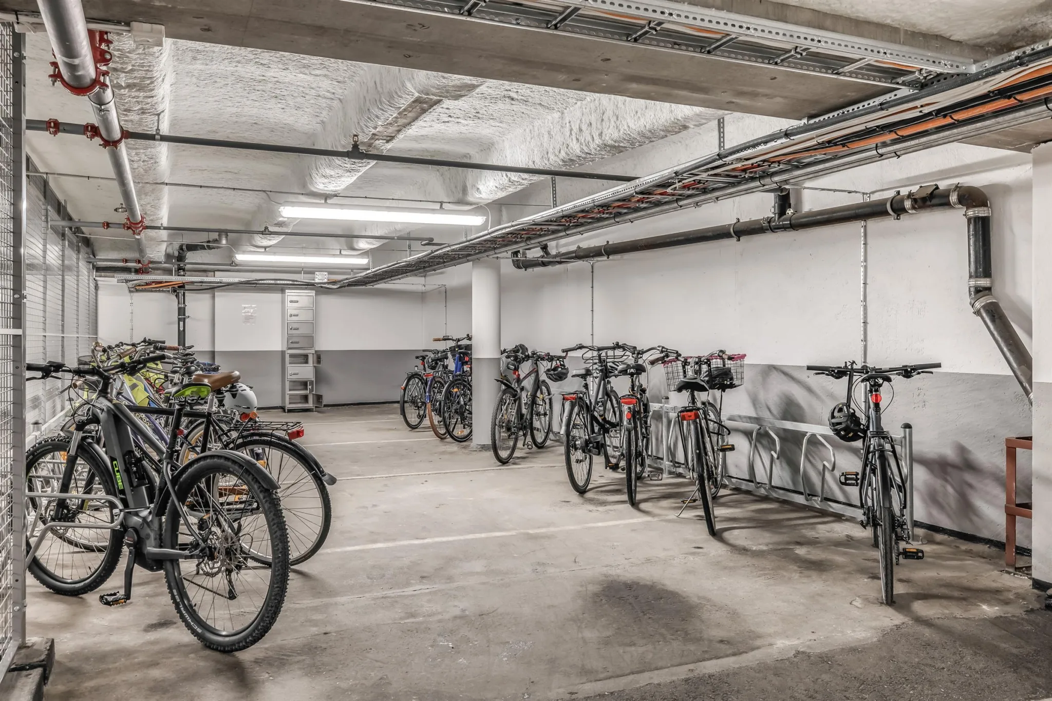 En cykelparkering inomhus med flera cyklar på rad i ett betongrum med rör i taket.
