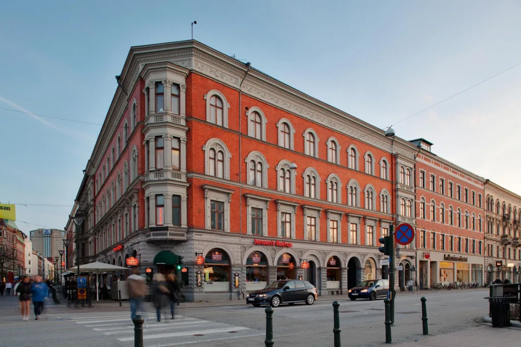 En röd tegelbyggnad med stora välvda fönster och butiker i bottenplan vid en korsning med bilar och fotgängare.