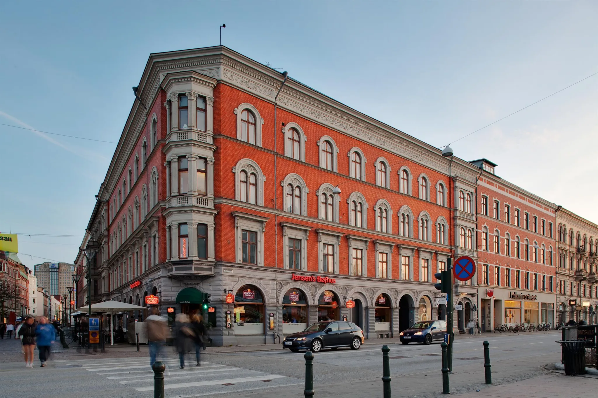 En röd tegelbyggnad med stora välvda fönster och butiker i bottenplan vid en korsning med bilar och fotgängare.