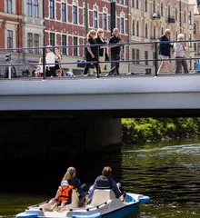 En bro med människor som går ovanpå, och under bron syns personer som paddlar en trampbåt på en kanal.