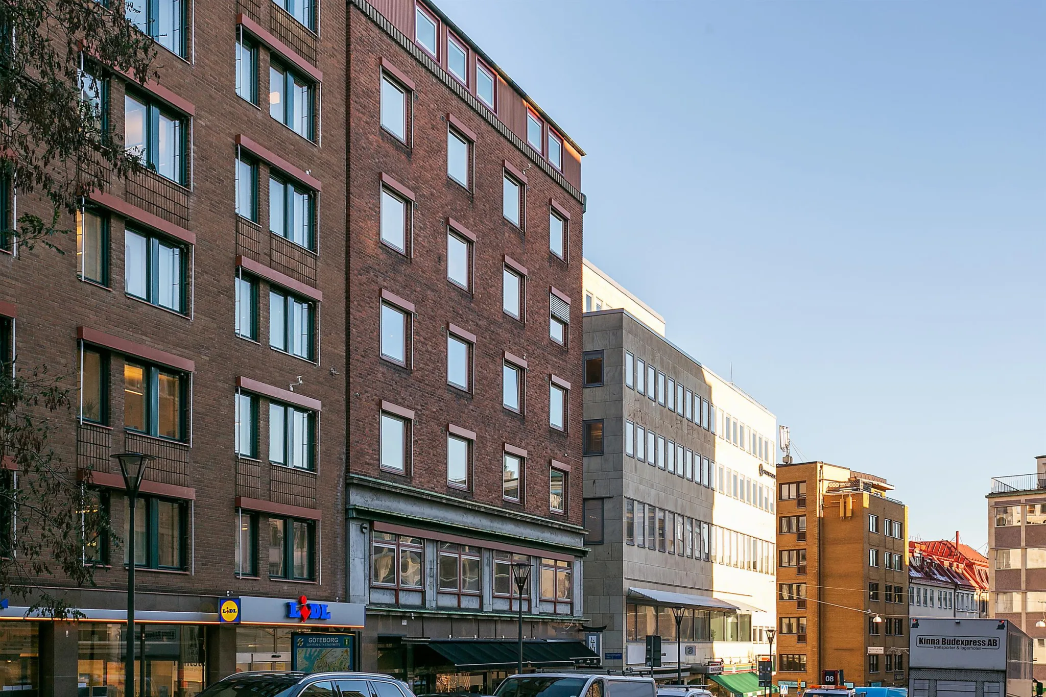 En gata med flervåningshus i olika arkitektonisk stil, parkerade bilar och klar blå himmel.