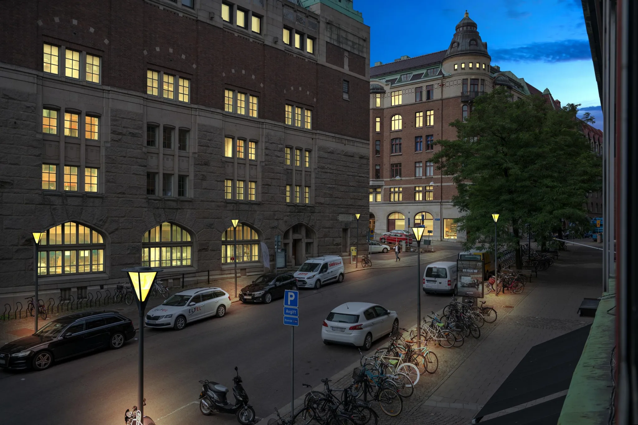 En kvällsbild av en upplyst gata med parkerade bilar, cyklar och äldre byggnader med tända fönster.