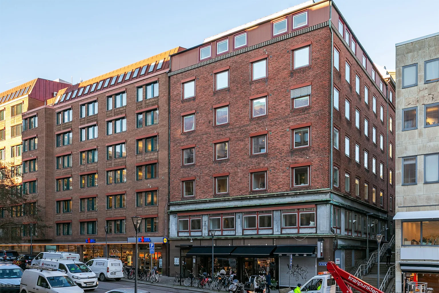 En tegelbyggnad med flera våningar och stora fönster, omgiven av bilar och fotgängare.