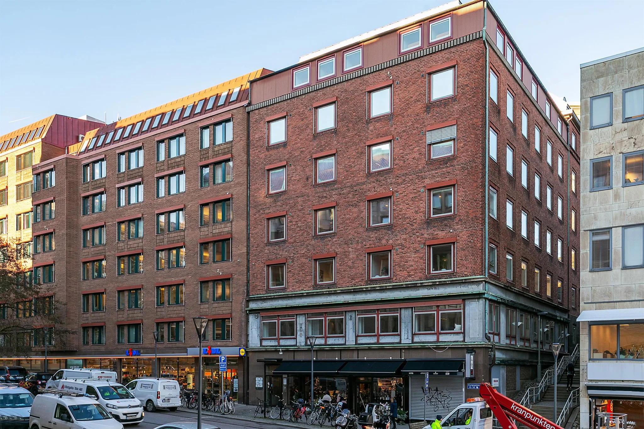 En tegelbyggnad med flera våningar och stora fönster, omgiven av bilar och fotgängare.