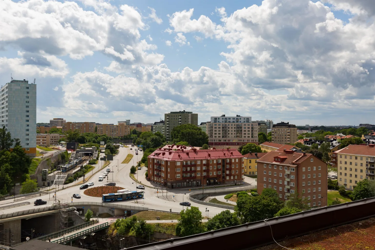 Utsikt över ett bostadsområde med röda tak, gröna träd och en molnig himmel.