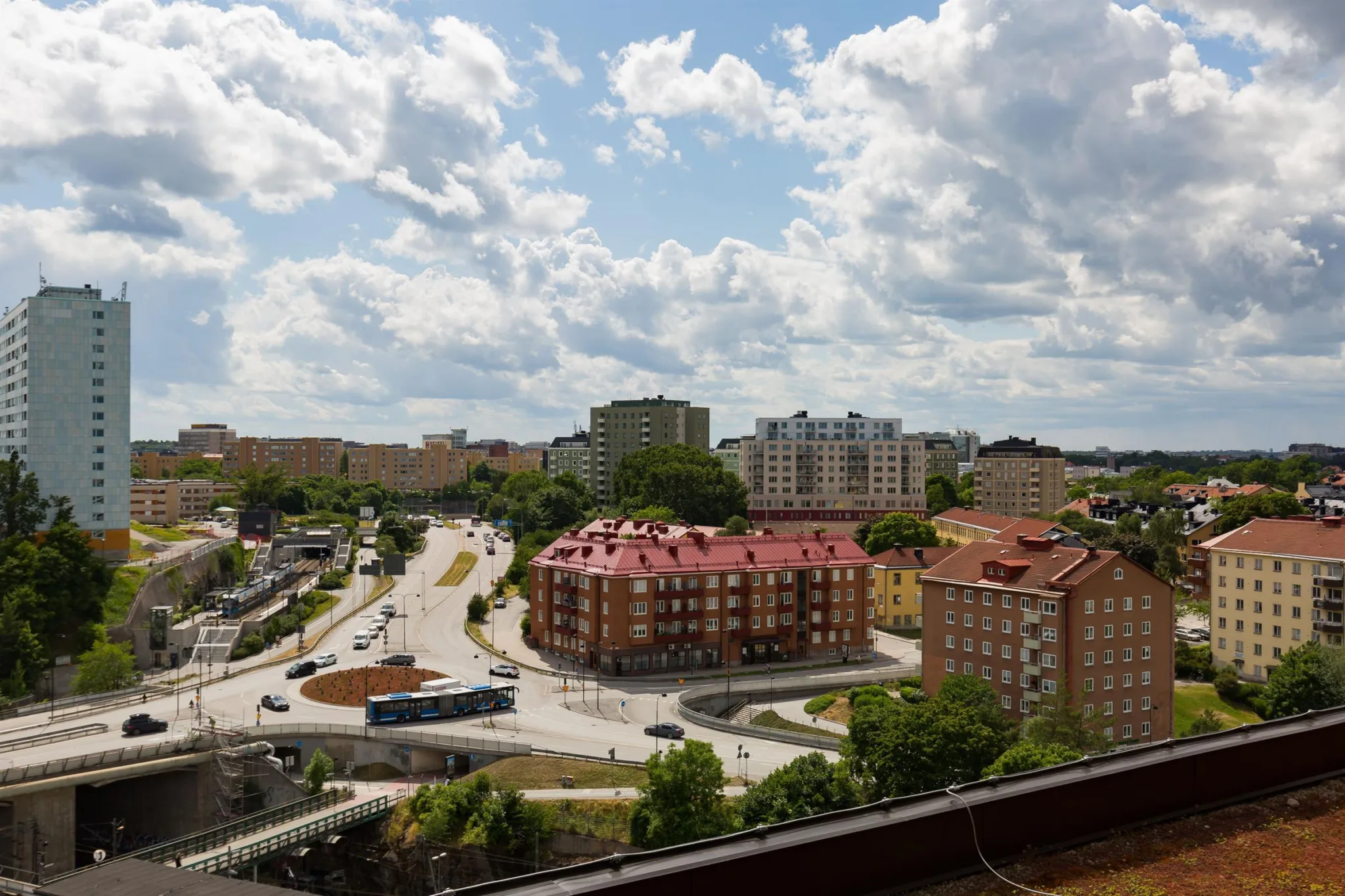 Utsikt över ett bostadsområde med röda tak, gröna träd och en molnig himmel.