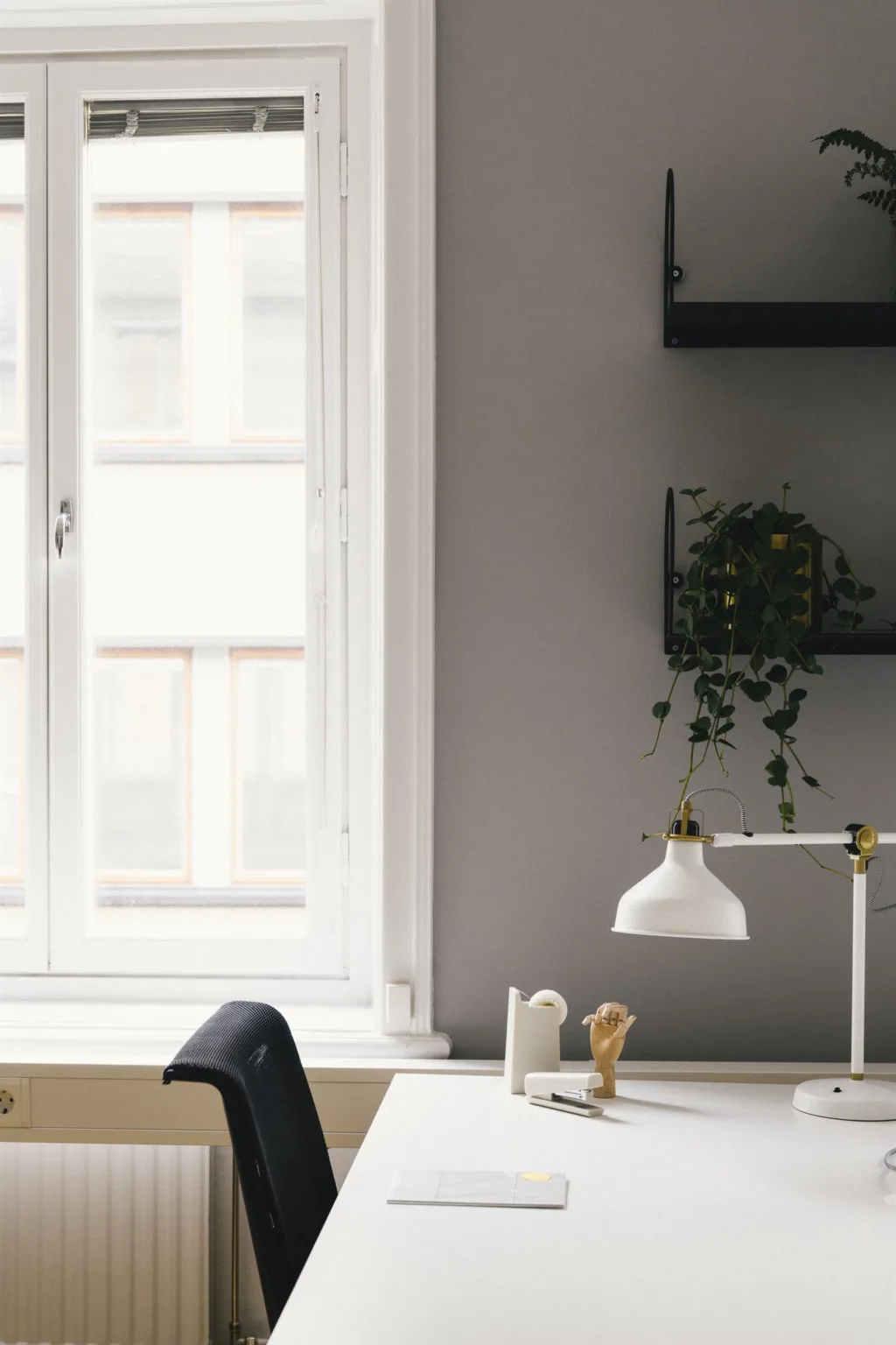 En skrivbordsyta med en vit lampa, kontorsmaterial och en växt bredvid ett stort fönster.