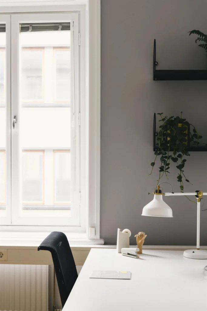 En skrivbordsyta med en vit lampa, kontorsmaterial och en växt bredvid ett stort fönster.