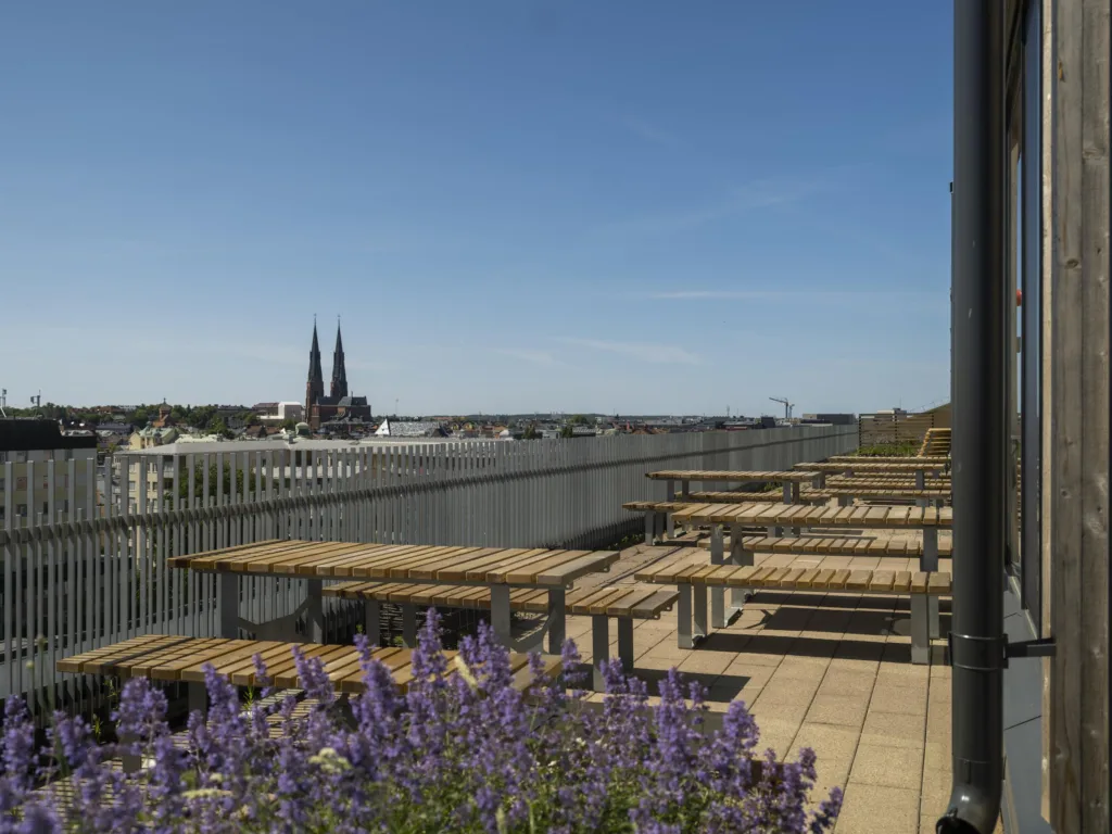 4. En takterrass med sittbänkar och bord, omgiven av blommor och med utsikt över en katedral i fjärran.