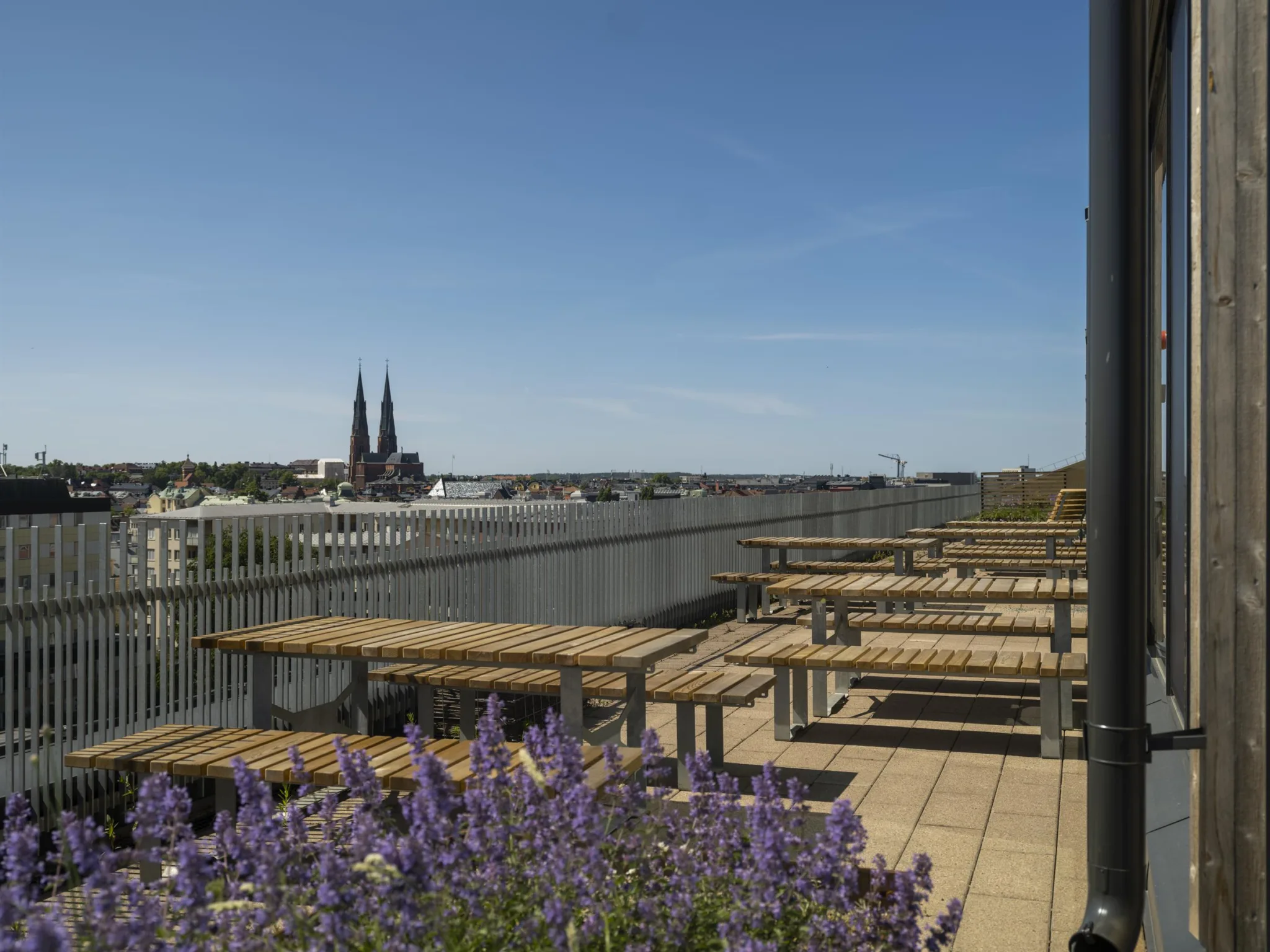 4. En takterrass med sittbänkar och bord, omgiven av blommor och med utsikt över en katedral i fjärran.
