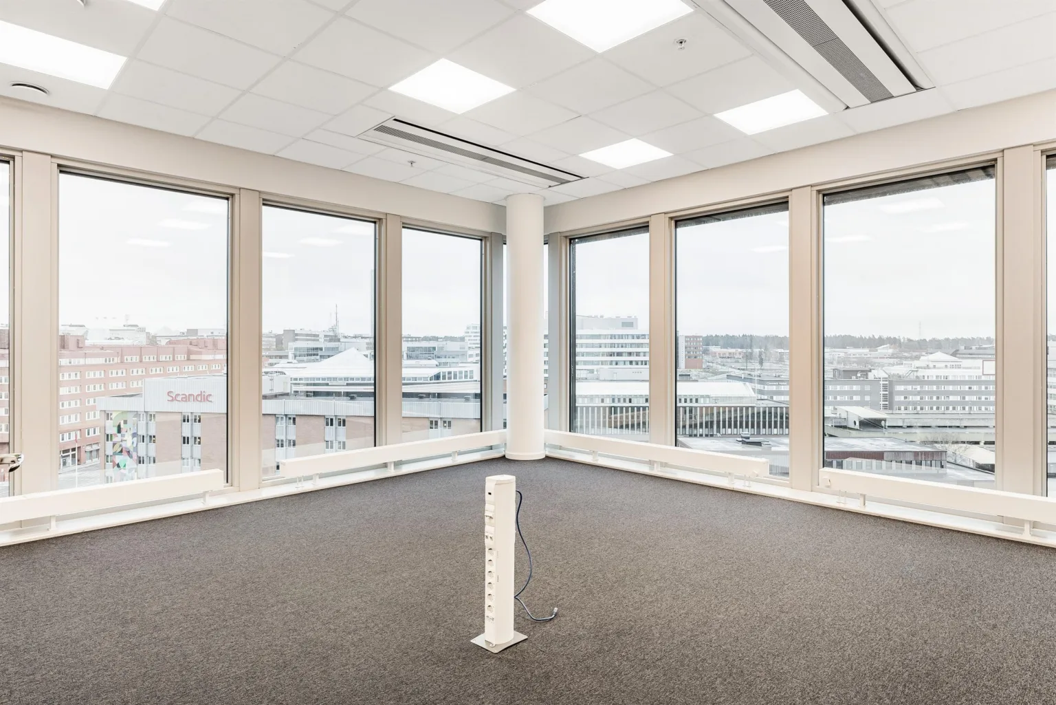 5. Ett hörnkontor med beige matta, stora fönster som ger utsikt över stadsmiljön och vita väggar.