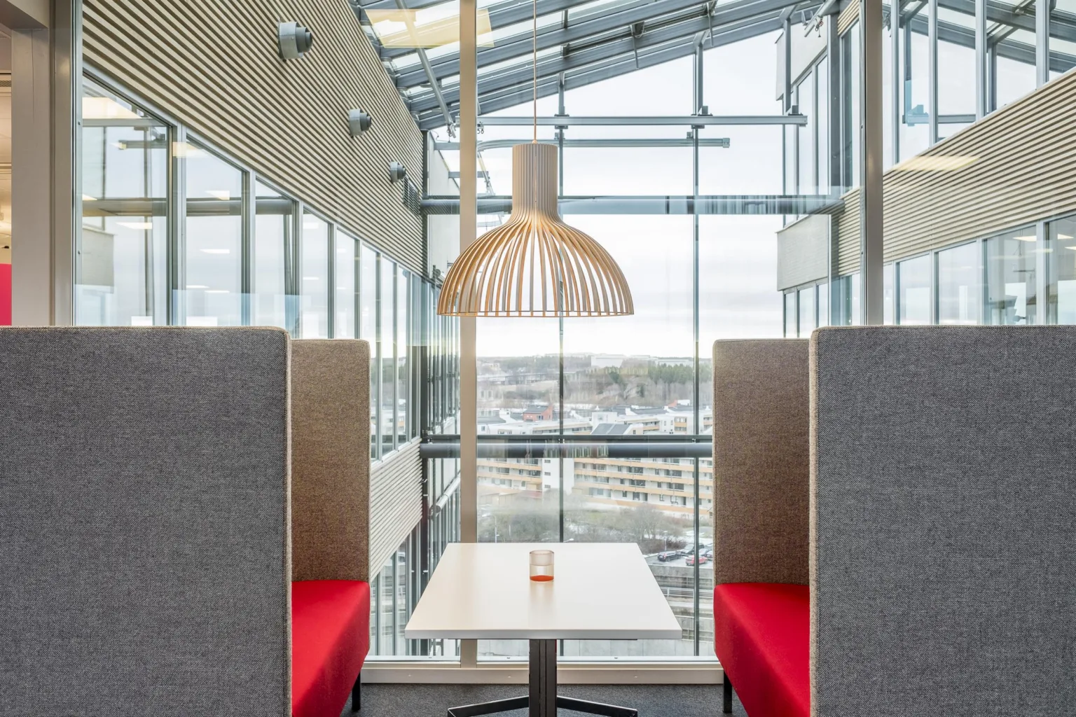 2. Ett modernt sittområde med två gråa bås med röda dynor, ett litet bord emellan och en träinspirerad taklampa ovanför, med stora fönster som visar en utsikt över staden.