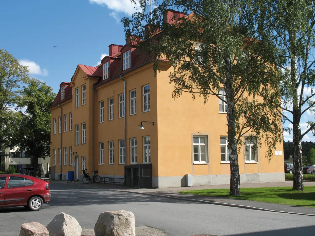 En gul byggnad med röda tak och fönster, omgiven av träd och en parkerad bil.