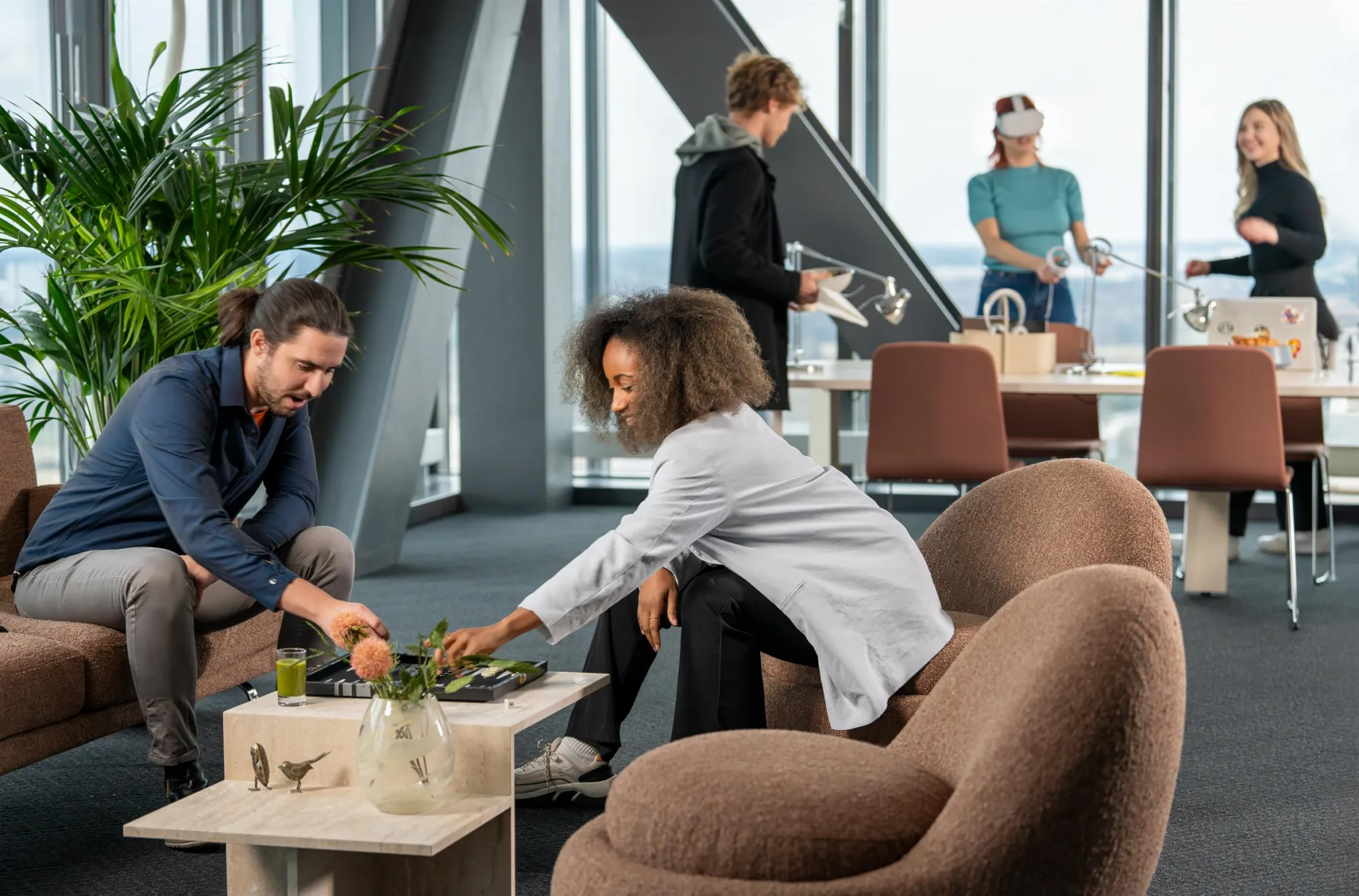 4. Två personer sitter vid ett bord och arrangerar blommor, medan andra interagerar i bakgrunden i ett kontorsutrymme.