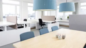 3. Ett kontorsutrymme med skrivbord, kontorsstolar, stora blå taklampor och ett mötesbord i förgrunden.