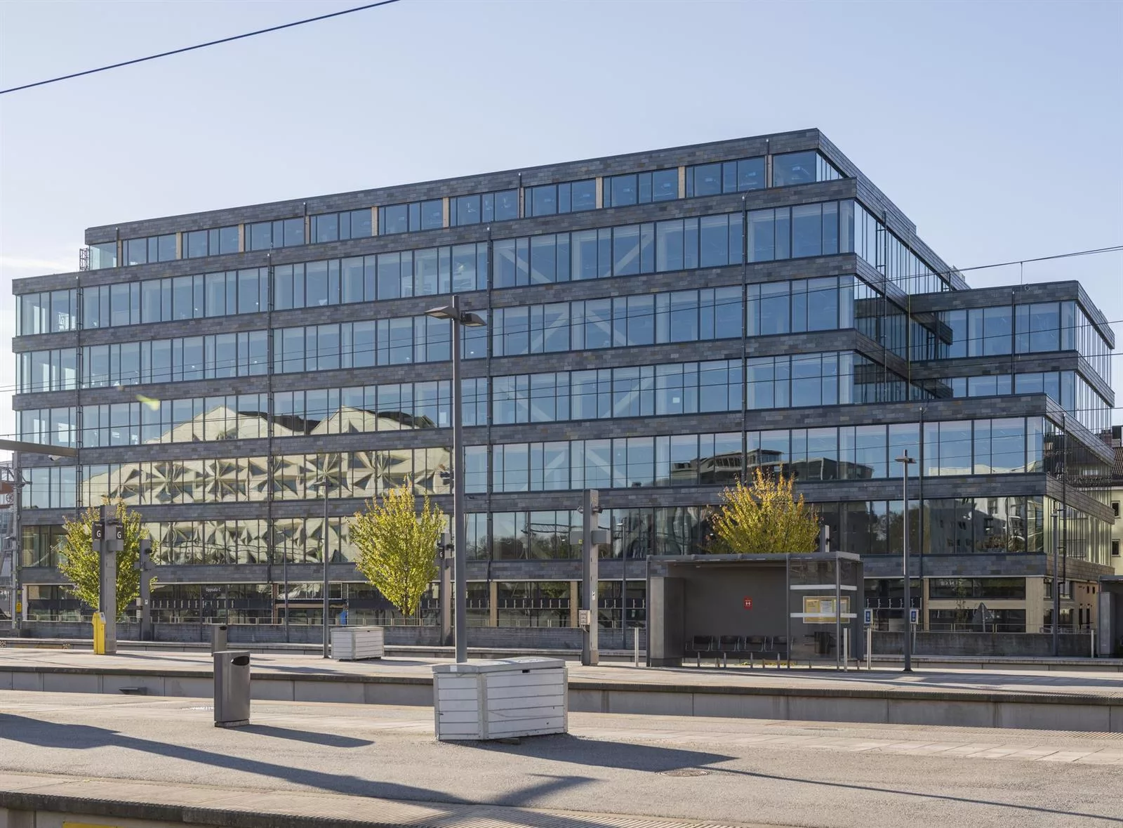 En modern glasbyggnad med reflekterande fasad, omgiven av en öppen gångväg och växtlighet.